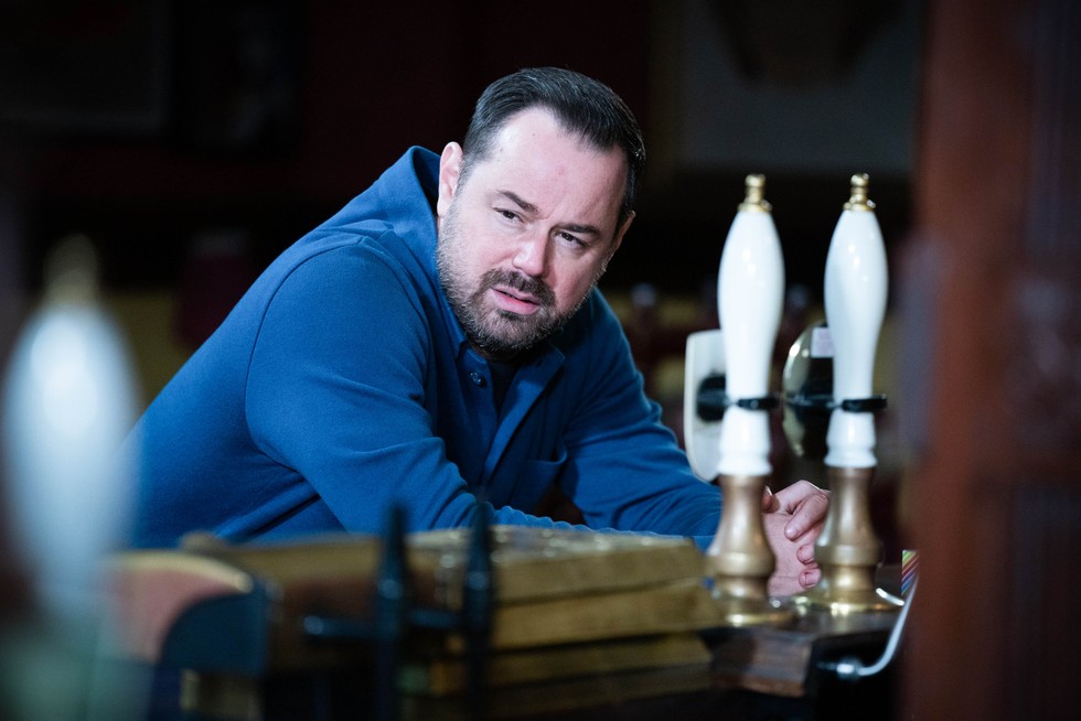 Danny Dyer as Mick Carter in EastEnders, leaning on the bar at the Queen Vic with beer pump handles in the foreground. He is wearing a blue shirt and looks upset.